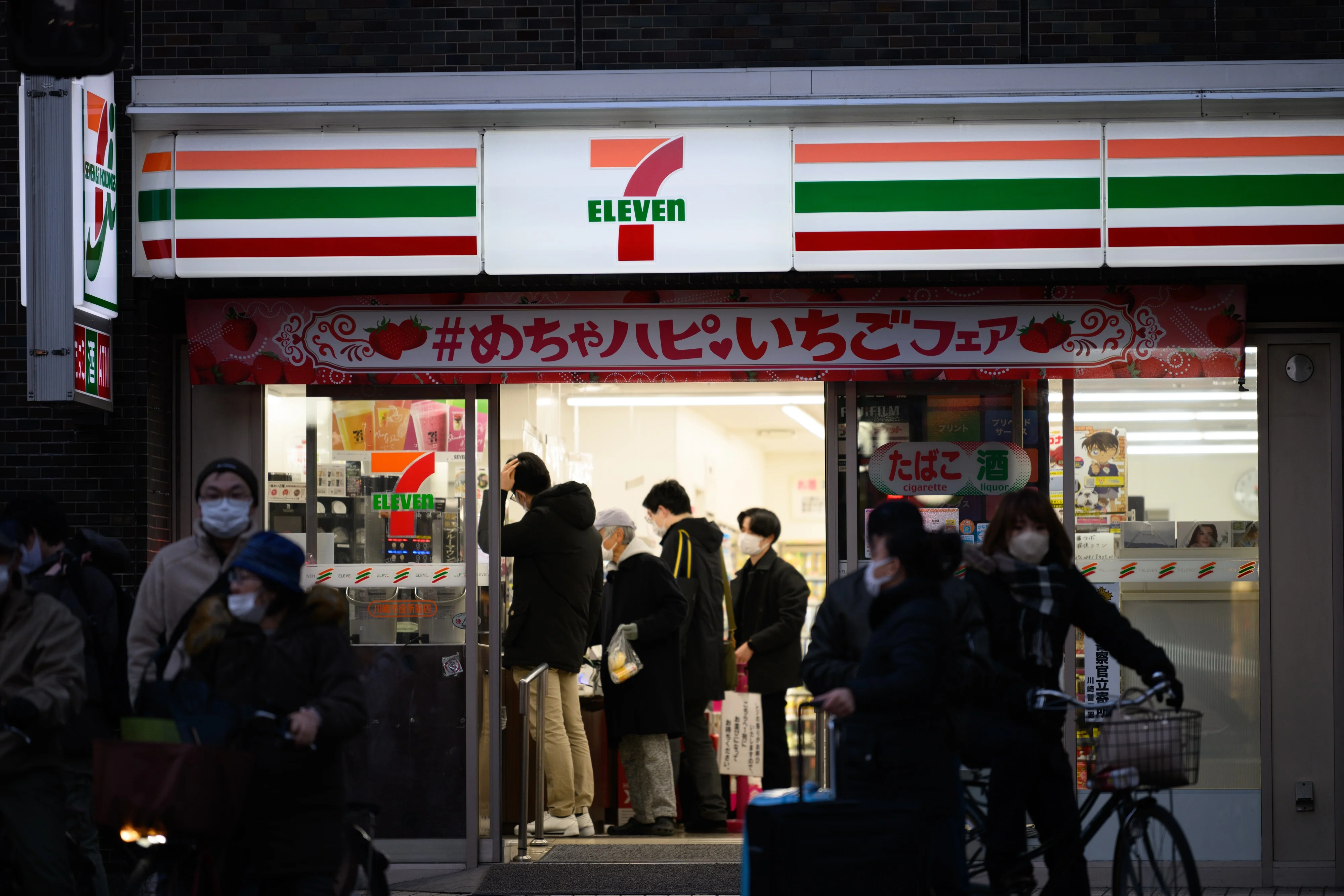 Fachada noturna de uma loja de conveniência 7-Eleven, com o logotipo brilhando em vermelho, laranja e verde. Há pessoas entrando e saindo, e veículos estacionados. A cena transmite um ambiente urbano movimentado e a presença da marca.