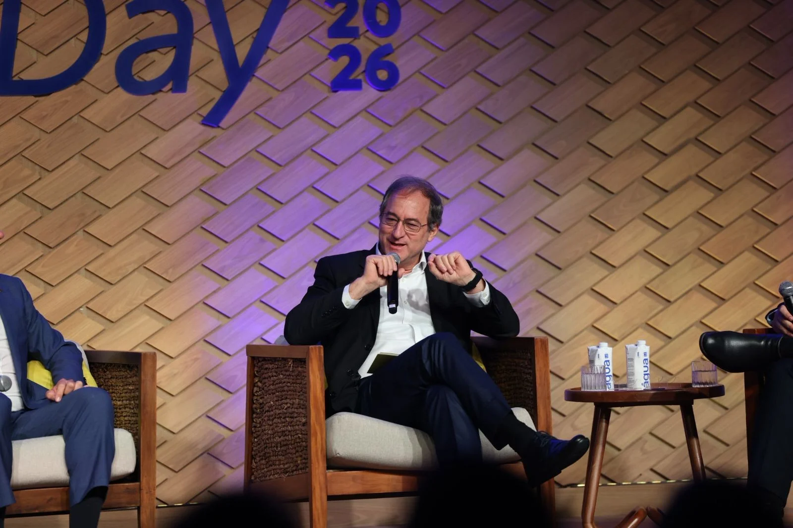 Executivo brasileiro de meia-idade em terno azul escuro gesticulando durante apresentação em evento corporativo, com gráficos financeiros digitais ao fundo.