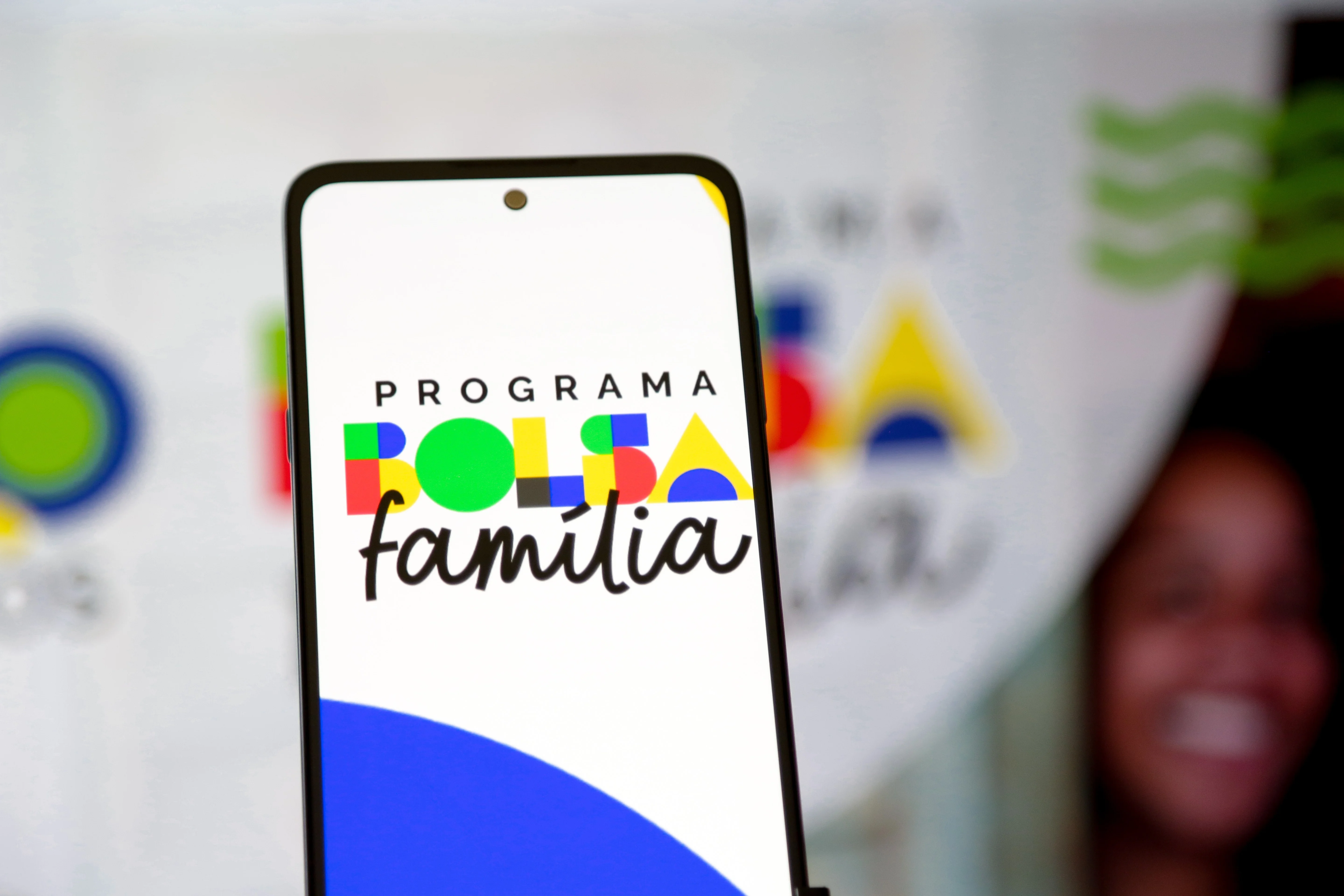 Mãos de uma pessoa segurando um cartão do Bolsa Família e um smartphone com o aplicativo Caixa TEM aberto, mostrando o saldo. Ao fundo, uma família de baixa renda sorrindo, simbolizando o impacto positivo do benefício.