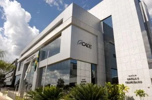 Uma mão segura um carimbo com o logo do CADE, simbolizando a aprovação ou reprovação de fusões e aquisições. Ao fundo, gráficos financeiros abstratos e uma balança da justiça embaçada, com cores sóbrias e iluminação dramática.