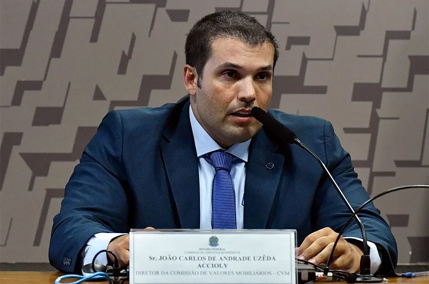 Um homem de cabelos grisalhos, em terno escuro, gesticula em um pódio com o logo da CVM ao fundo, representando uma autoridade financeira em discurso. A imagem transmite seriedade e foco.