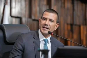 Homem jovem de terno escuro e gravata azul, com aparência de político brasileiro, discursando em um plenário legislativo com painéis de madeira ao fundo.