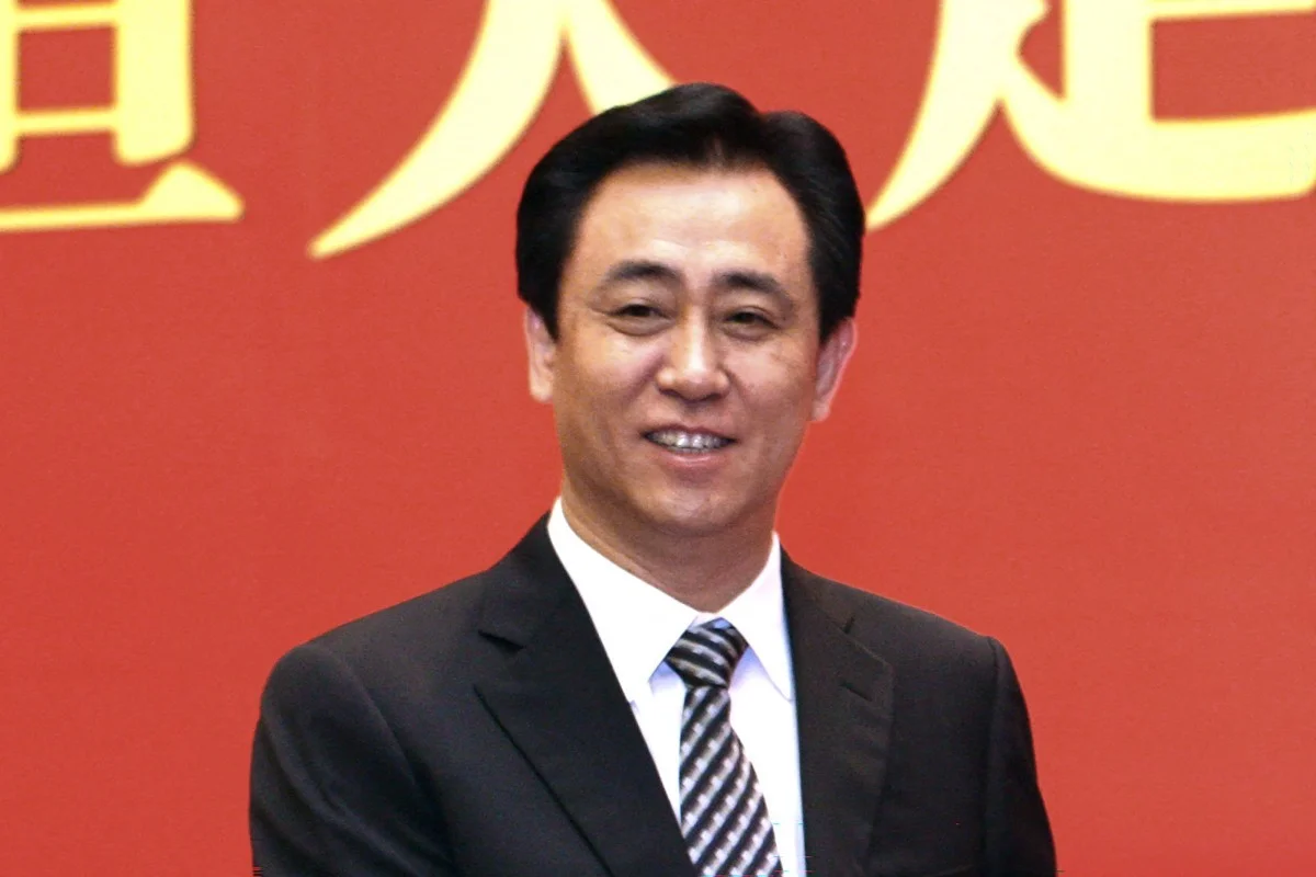O ex-presidente da Evergrande, Xu Jiayin, um homem de meia-idade com óculos e expressão séria, sentado formalmente. Ele usa um terno escuro com gravata e está em um ambiente corporativo com iluminação suave, transmitindo uma atmosfera de seriedade e forma