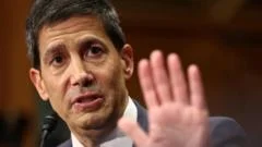 Kevin Warsh, um homem de meia-idade com cabelos grisalhos e terno escuro, gesticula enquanto fala em uma audiência no Senado dos EUA, com senadores ao fundo. O ambiente é formal e tenso, com iluminação dramática.