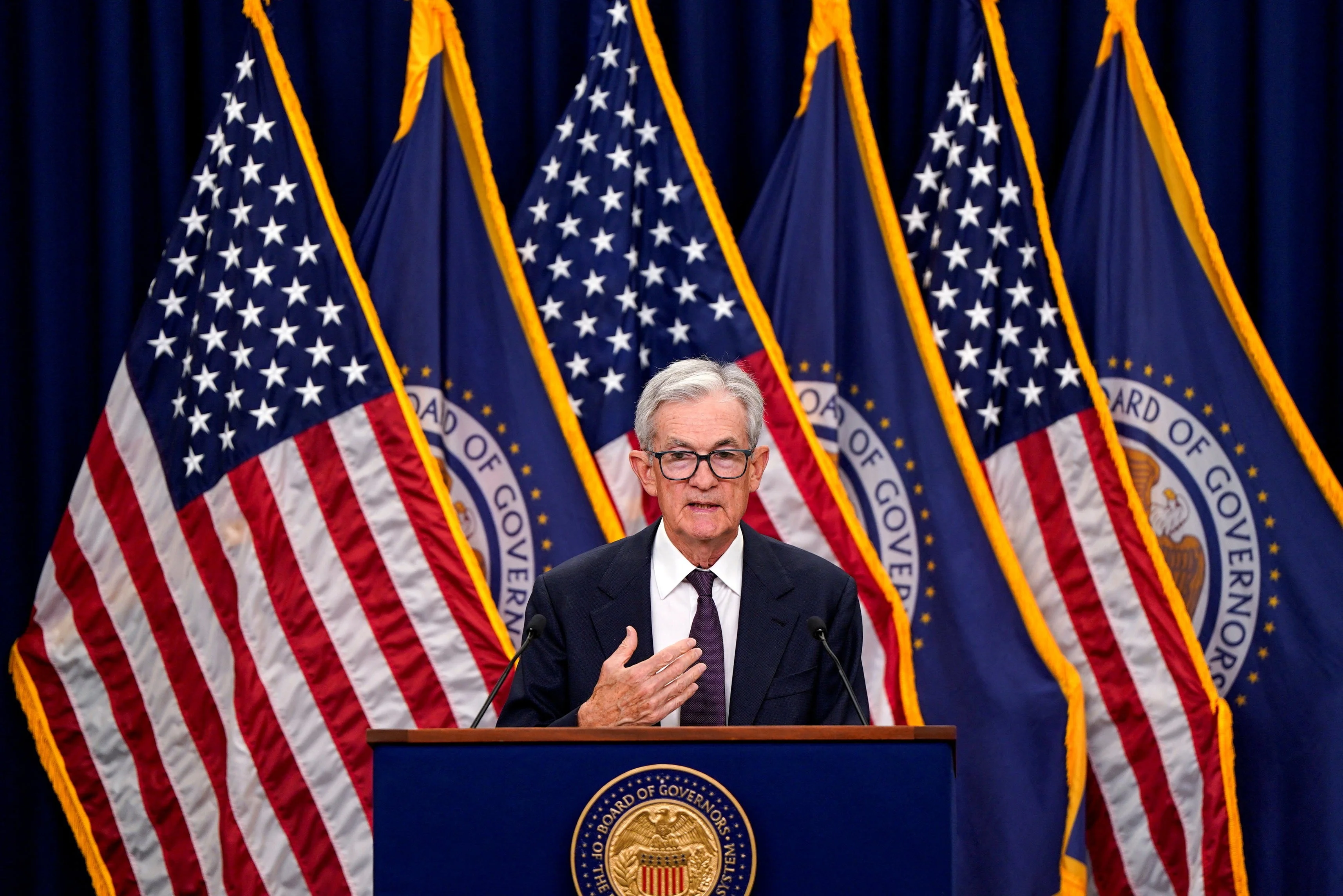 Um homem idoso de cabelos brancos e terno escuro, representando o presidente do Fed, em um púlpito. Ao fundo, o imponente prédio do Federal Reserve e gráficos financeiros digitais sobrepostos, sob iluminação dramática.