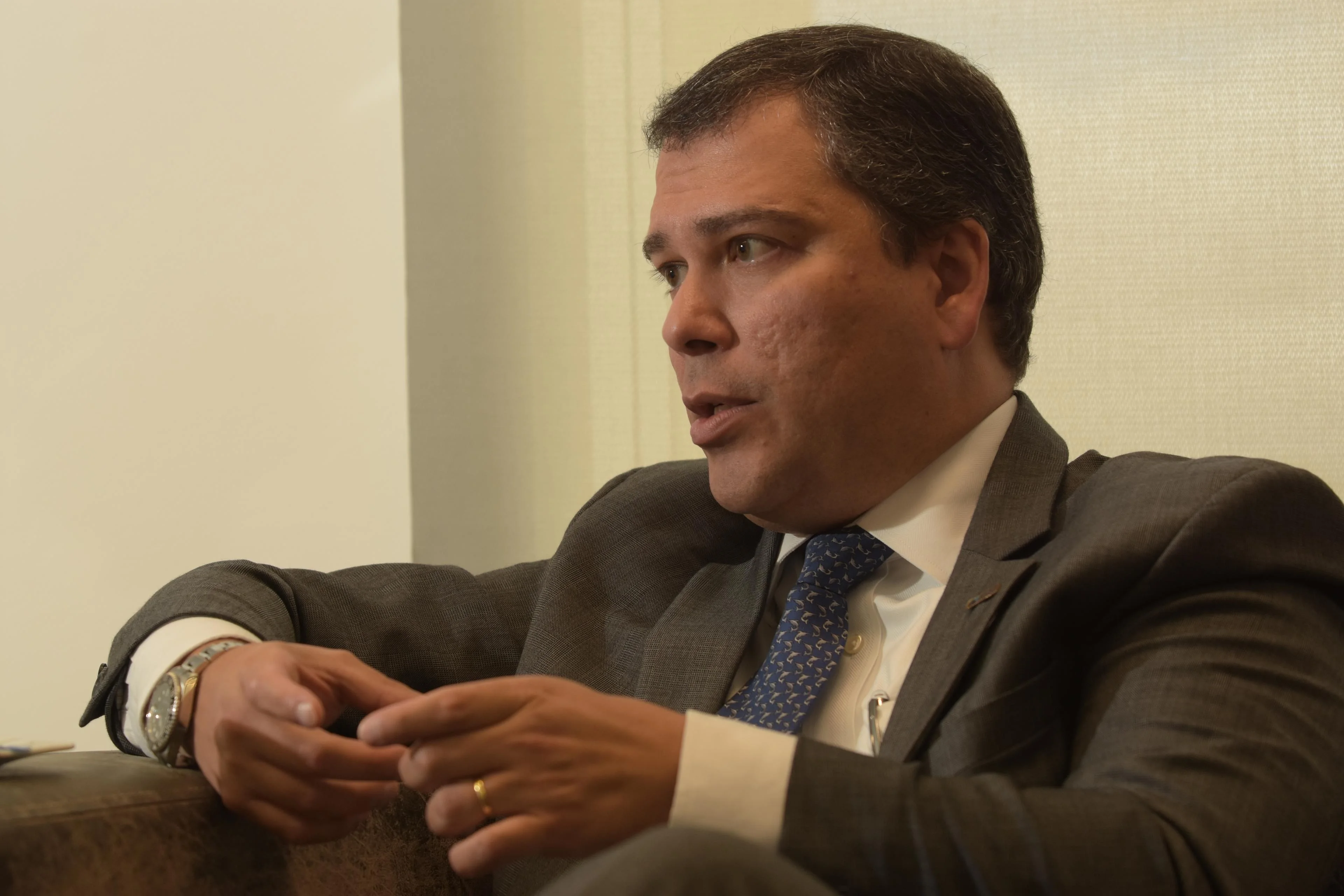 Uma mão com um anel formal, vestindo um terno escuro, está segurando um tablet que exibe gráficos financeiros em tons de verde e vermelho, com um fundo desfocado de escritórios corporativos e símbolos de dólar. A cena sugere investigação e corrupção no se