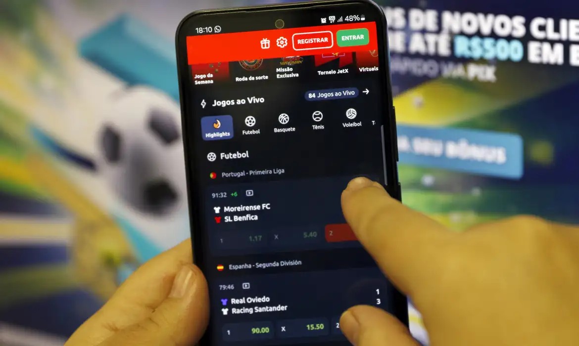 Uma mão digital simbolizando o governo bloqueia o acesso a um site de apostas online exibido em um smartphone, com moedas e símbolos do Bolsa Família e BPC ao fundo, em um cenário de proteção financeira.