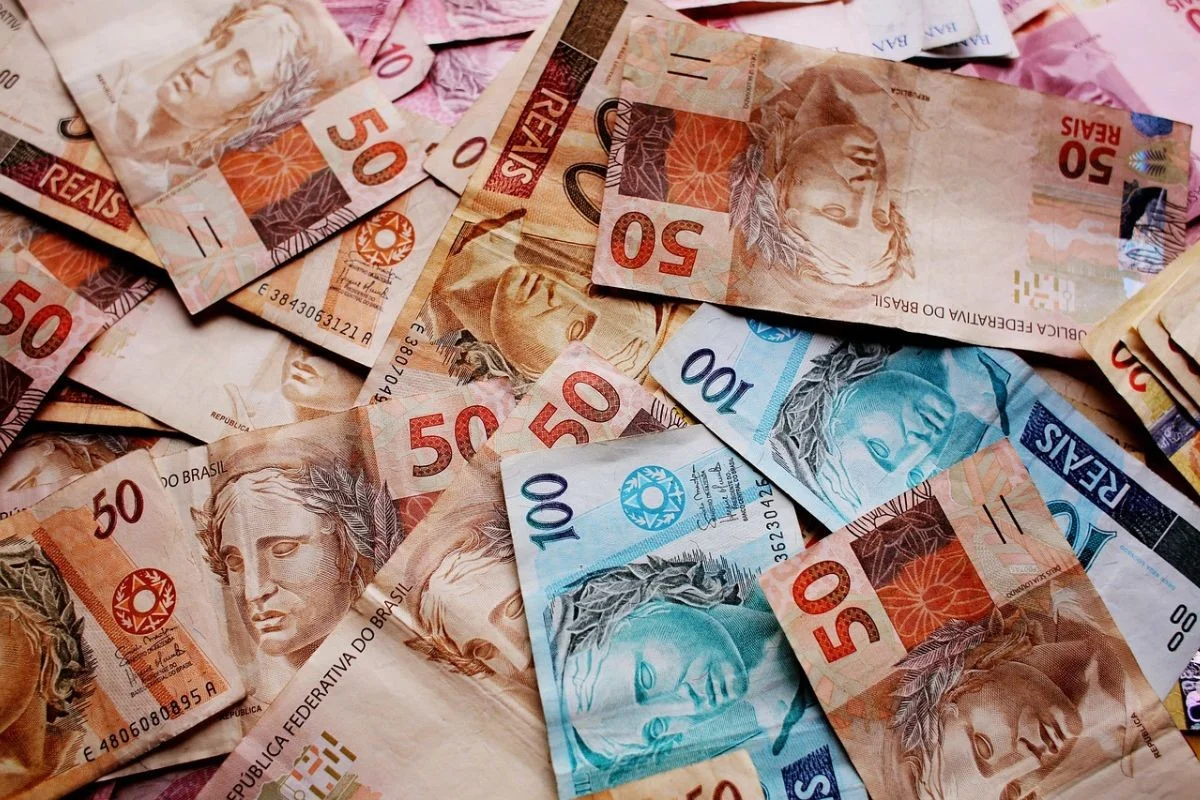 Close-up de notas de 100 reais empilhadas ao lado de um tablet exibindo gráficos financeiros de barras e linhas em tons de azul e verde, ambiente de escritório moderno.