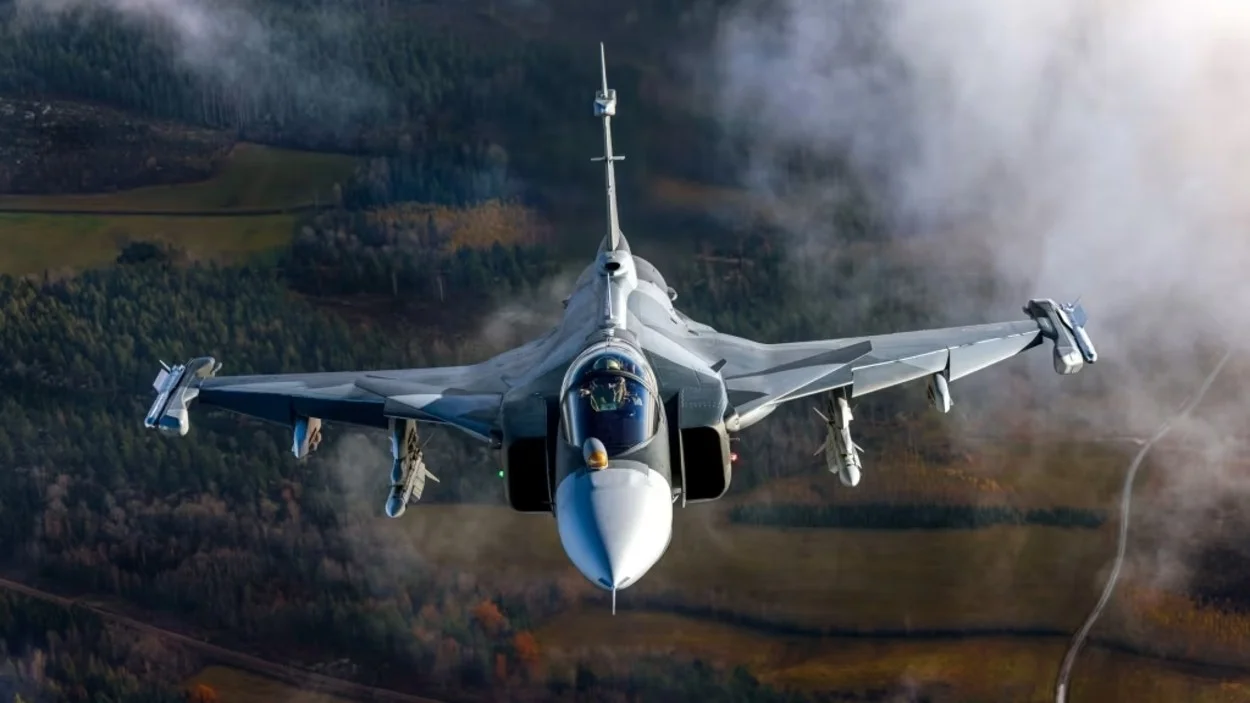 Caça Gripen F-39 cinza-escuro em voo dinâmico sobre um céu azul com nuvens. A aeronave exibe seu design aerodinâmico e tecnologia avançada, com detalhes vermelhos e azuis, transmitindo poder e agilidade.