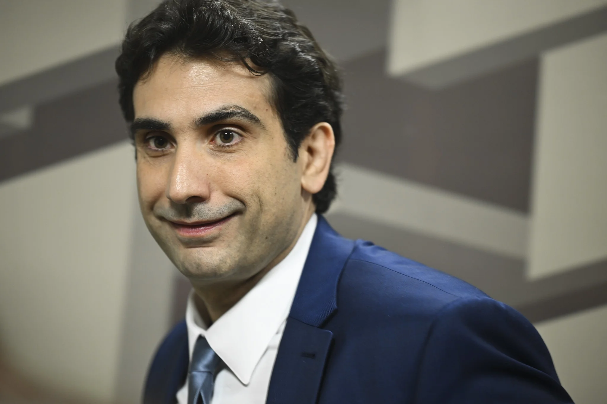 Um homem de meia-idade, cabelos grisalhos, terno escuro, gesticulando em um pódio com microfones. Ao lado, uma jabuticabeira frutífera com cartões de crédito pendurados nos galhos, simbolizando o sistema financeiro único do Brasil.