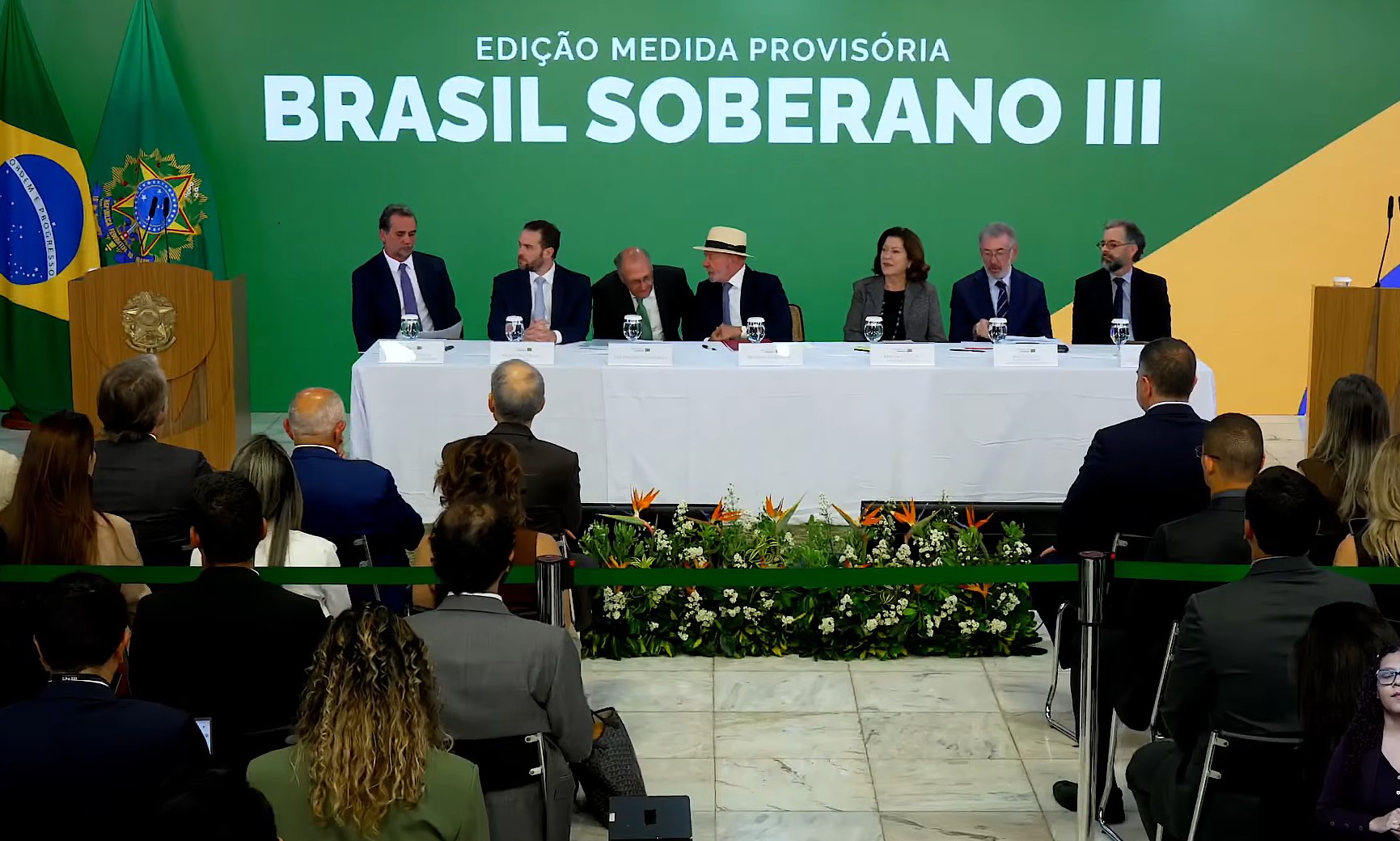Um presidente brasileiro idoso de cabelos brancos, em um paletó escuro, discursa em um pódio com o símbolo do governo, anunciando um pacote econômico. Ao fundo, telas exibem gráficos e símbolos de moedas, com foco em finanças e recuperação econômica.