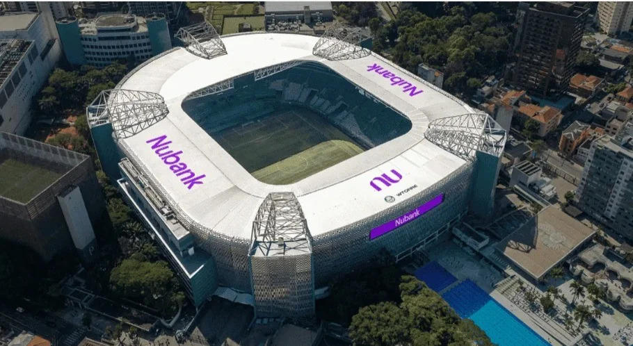 Visão aérea de um estádio de futebol ultra-moderno iluminado com luzes roxas neon ao entardecer, destacando a arquitetura contemporânea e uma atmosfera tecnológica.