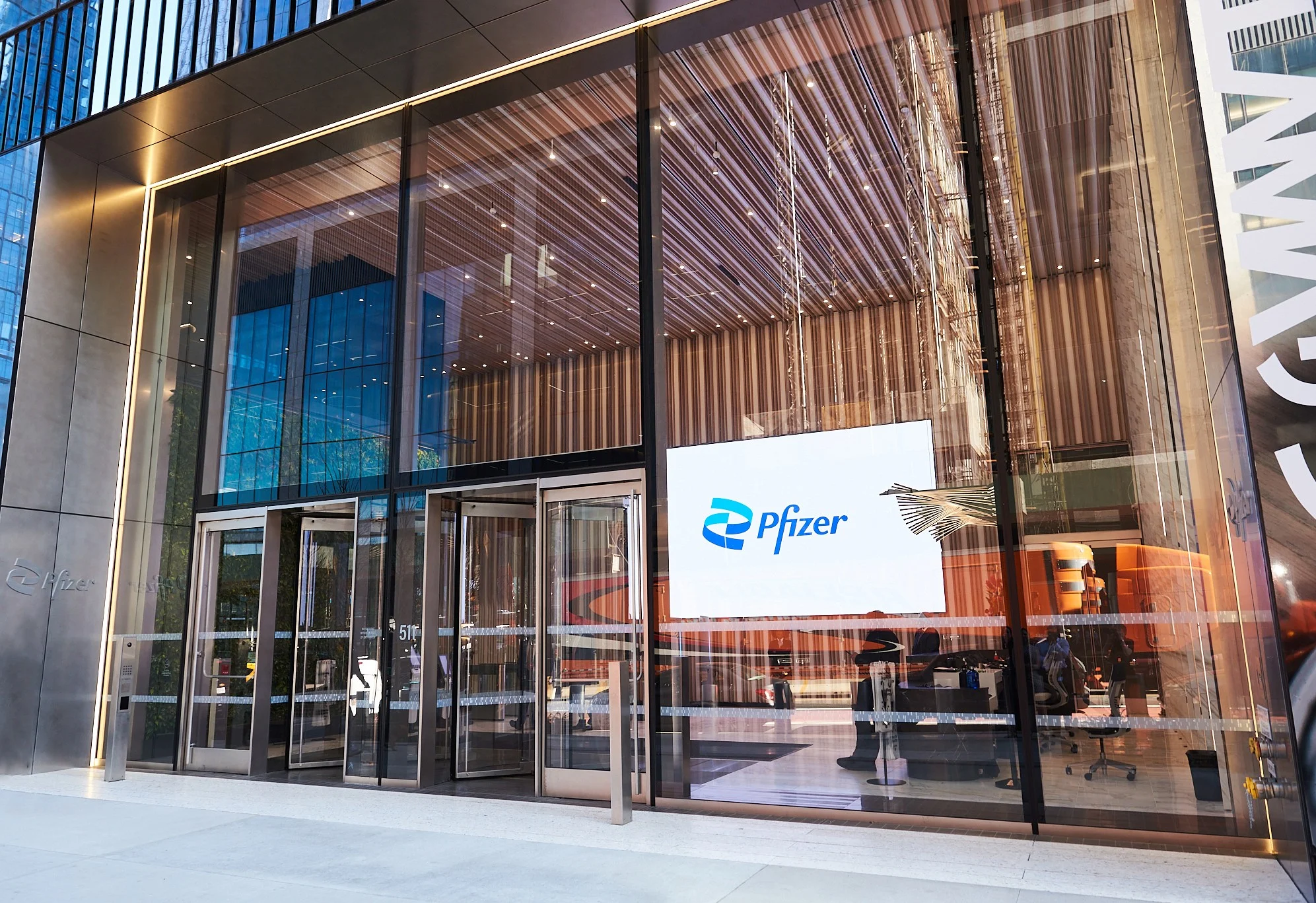Close-up de um frasco de medicamento moderno com o logotipo da Pfizer em foco seletivo, sobre uma mesa de escritório com gráficos financeiros ao fundo.
