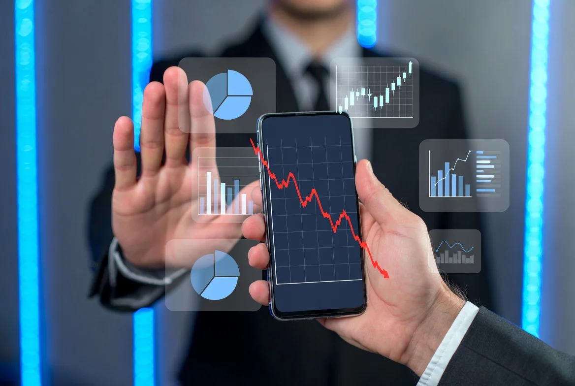 Uma mão masculina em terno segurando um smartphone que exibe um gráfico financeiro em queda, com a outra mão fazendo um gesto de 'pare' para um grupo de ícones de ações em segundo plano. O ambiente é um escritório moderno com luzes de LED azuladas.