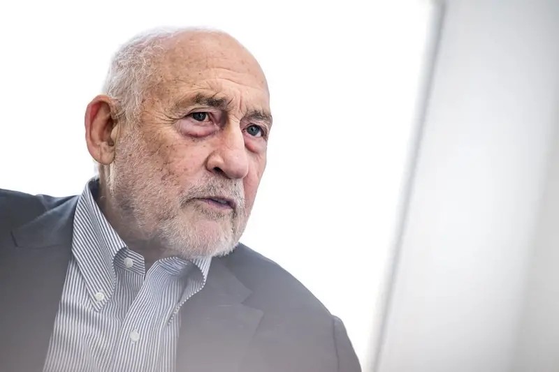 Um economista renomado, com cabelos brancos e óculos, em um púlpito ou mesa de entrevista, com um mapa-múndi digital e elementos do PIX ao fundo, simbolizando finanças globais e tecnologia.