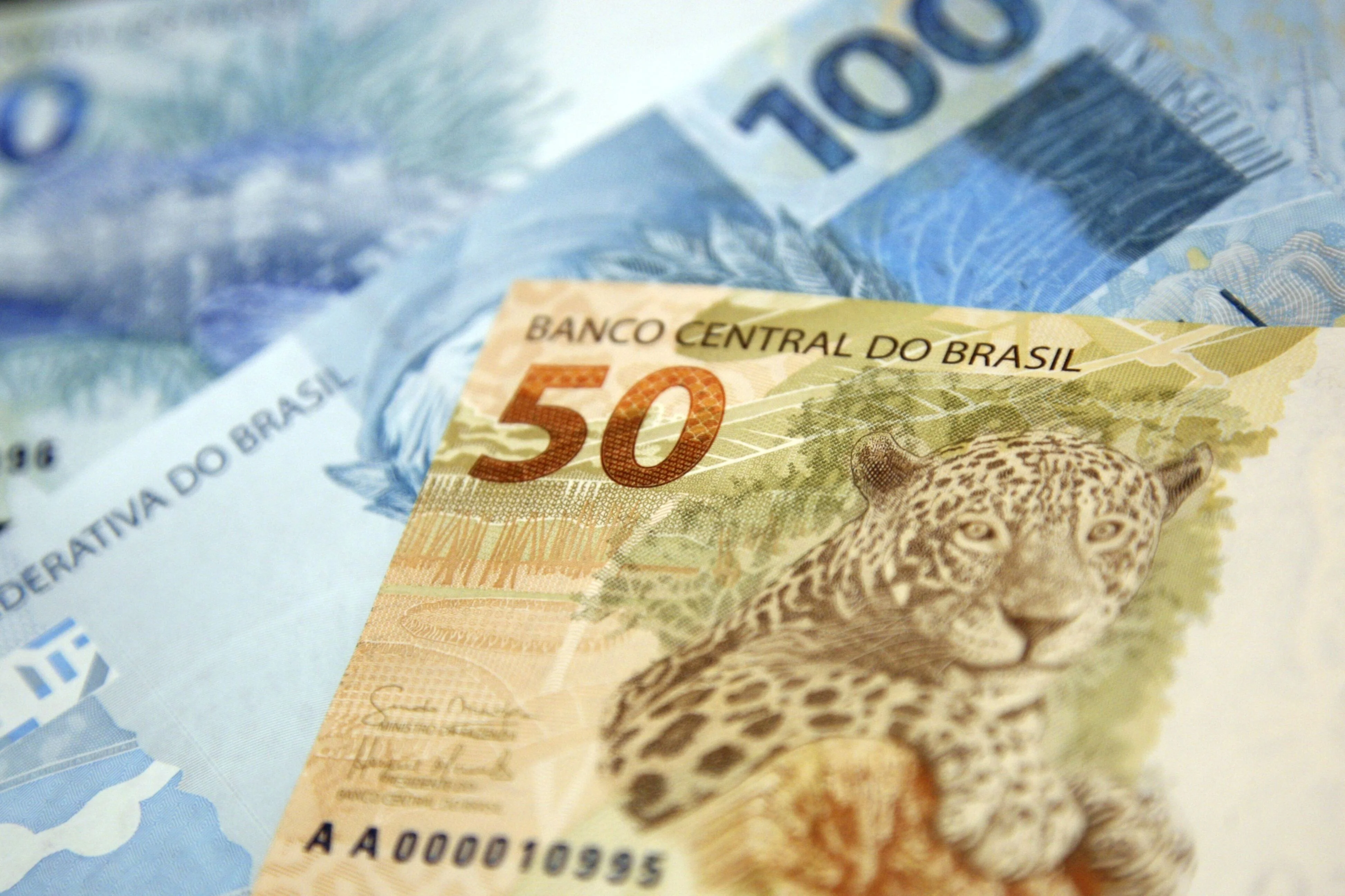 Mãos de um profissional da Receita Federal em uma mesa, analisando documentos fiscais e gráficos digitais em um tablet, com uma lupa e pilhas de dinheiro ao fundo. A cena transmite fiscalização e investigação tributária.