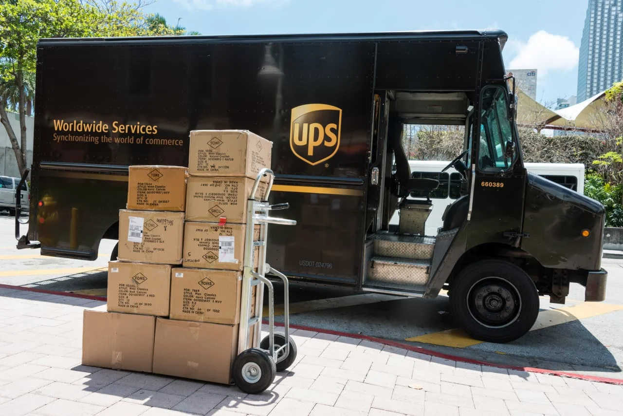 Uma van de entrega da UPS em movimento rápido, com um gráfico financeiro sobreposto mostrando uma queda acentuada. Moedas e uma calculadora estão em primeiro plano, simbolizando lucro e perda.