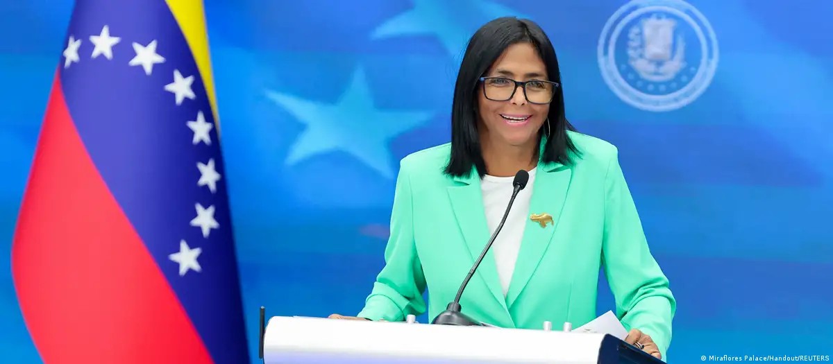 Uma mulher de meia-idade com cabelos escuros e roupas formais, identificada como a presidente interina da Venezuela, discursa em um pódio com microfones e bandeiras venezuelanas ao fundo, em um cenário de palácio. A imagem transmite seriedade e liderança 