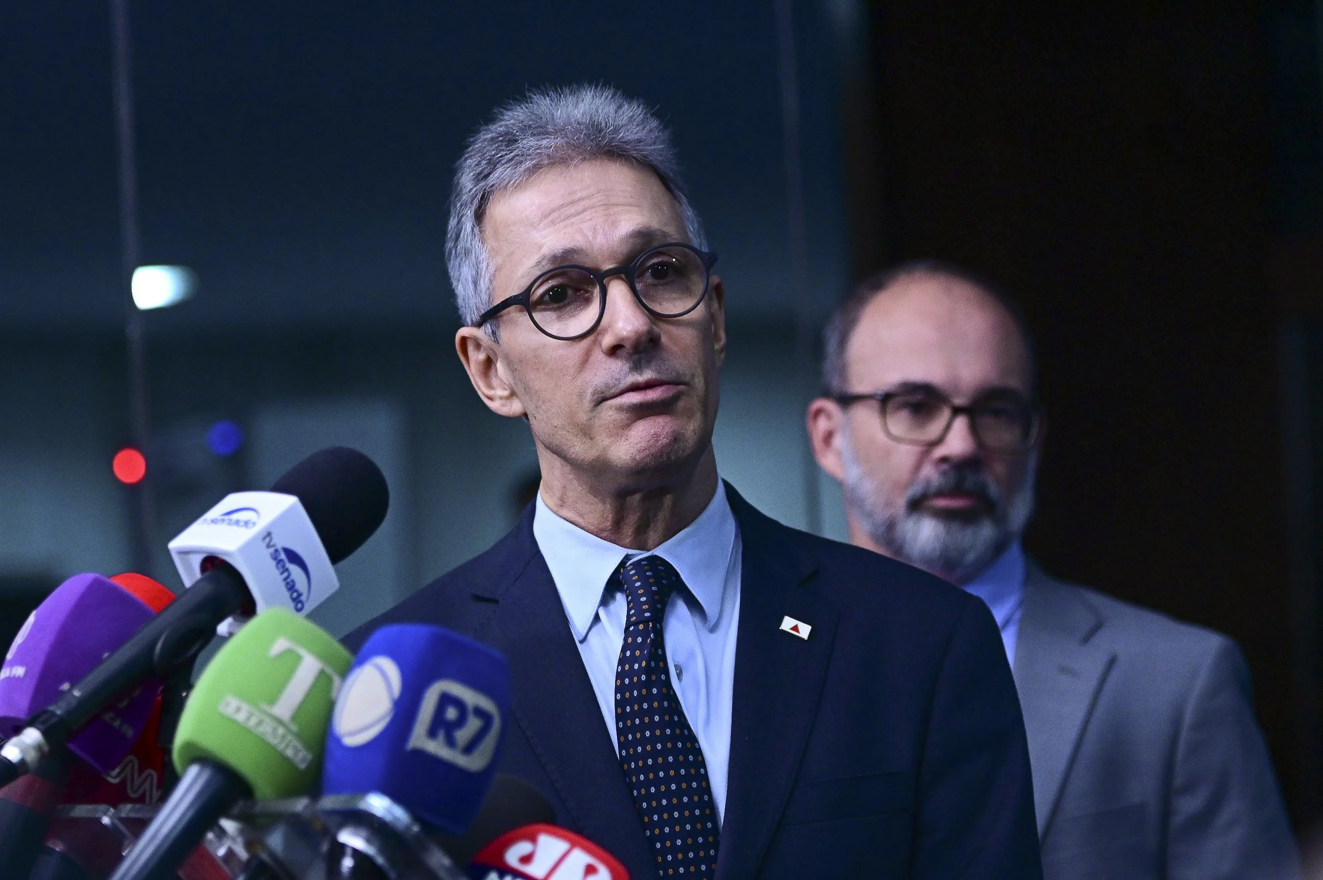 Um político de cabelos claros, em terno escuro, gesticulando com uma mão enquanto fala em um púlpito. O fundo está desfocado, indicando um evento público com a presença de uma plateia. A imagem transmite seriedade e debate.